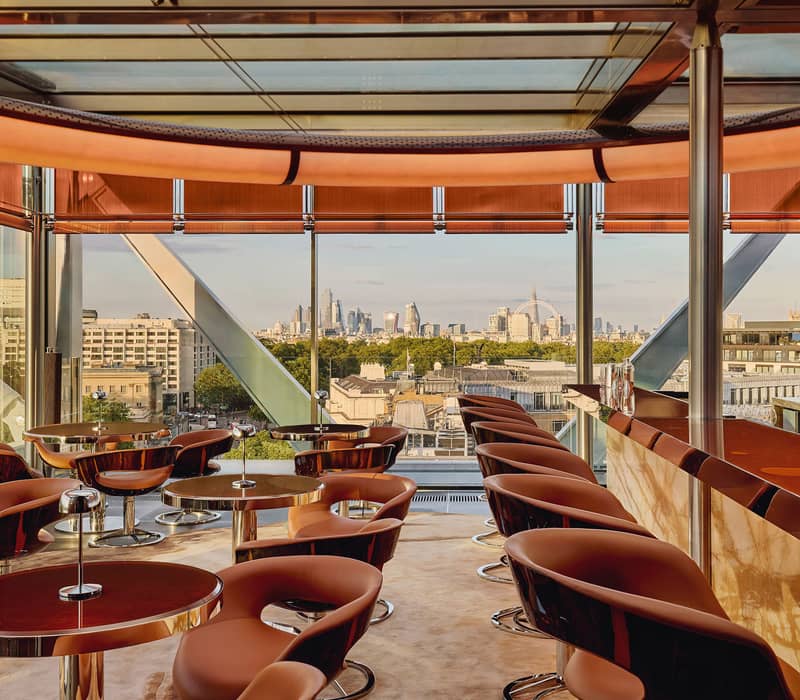 The Emory Rooftop bar, circular tables and brown leather chairs are placed in a bright room with views of the London skyline