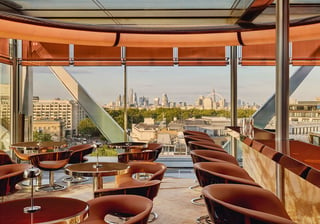 The Emory Rooftop bar, circular tables and brown leather chairs are placed in a bright room with views of the London skyline