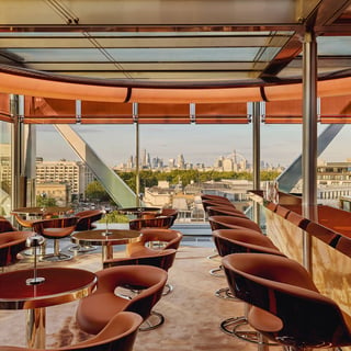 The Emory Rooftop bar, circular tables and brown leather chairs are placed in a bright room with views of the London skyline