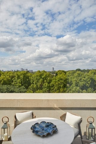 A round white table with a blue ceramic on the terrace with two arm chairs and white cushions overlooking Hyde Park