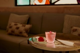 A pink cocktail sits on a glass table. A sofa and cushions are in the background