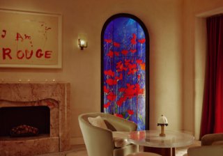 The Connaught Red Room. A round glass table with a small lamp sits to the right. Two beige arm chairs sit either side. In the background there is a blue and red flower art piece. To the left is a marble fireplace